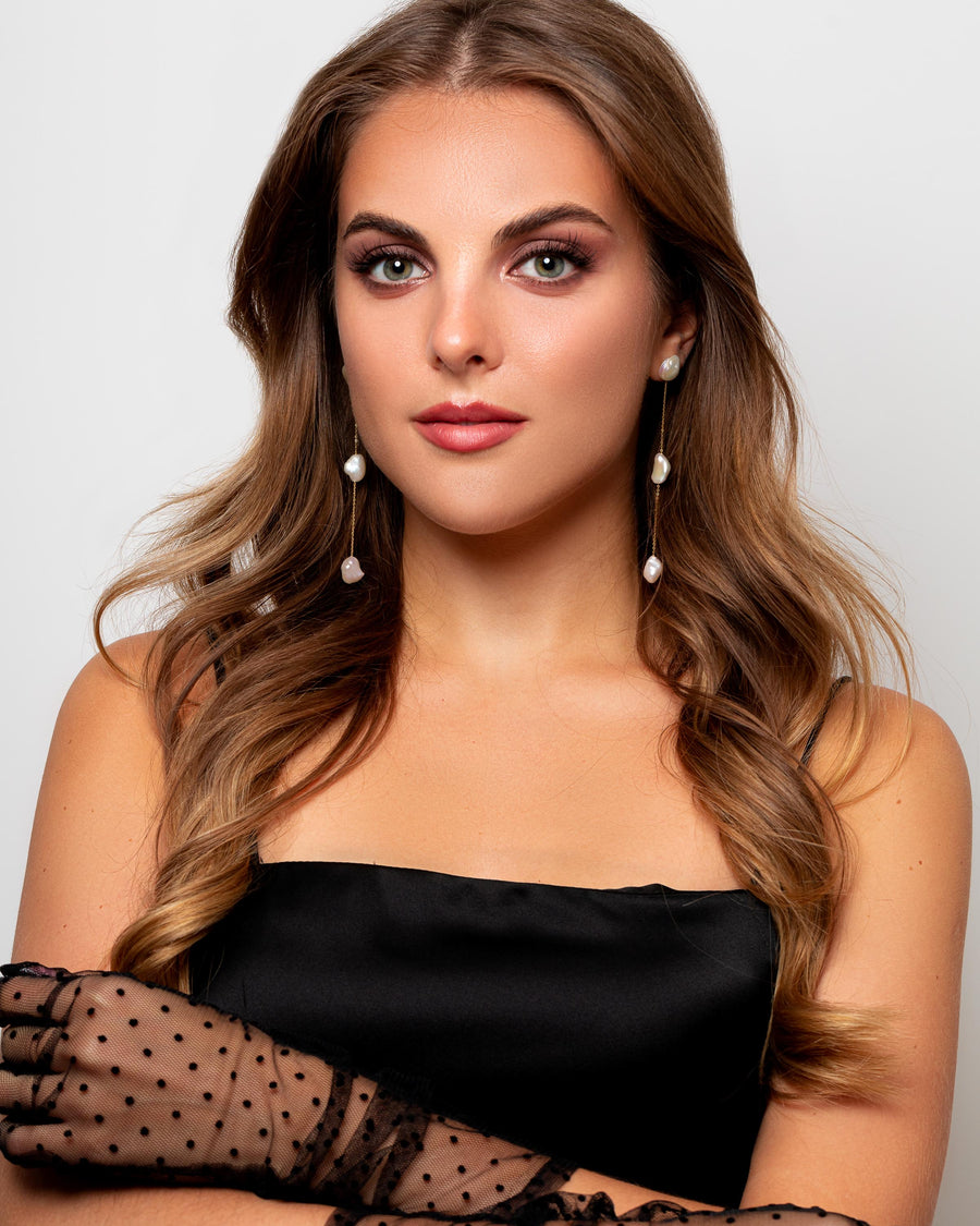 A Caucasian woman with brown hair wears elegant gold studs earrings with Baroque pearls