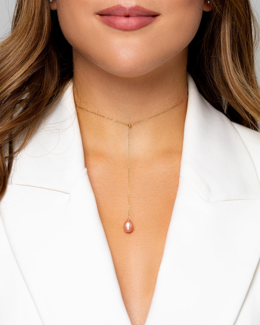 A beautiful gold necklace with a stunning pink teardrop pearl sits on a woman’s neckline